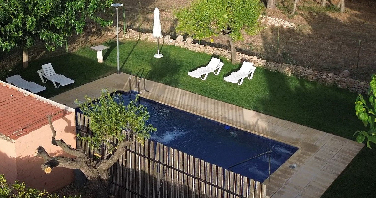 A private pool area with a blue swimming pool, white lounge chairs, a wooden fence, green lawn, and tall trees under a clear blue sky.